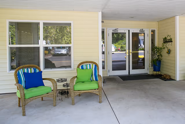 Covered entrance area of a facility with two wicker chairs featuring green cushions and blue pillows, a small black metal plant stand between them, a window with blinds, double glass doors with gold handles, a welcome sign on the wall, and a potted plant in a blue container.