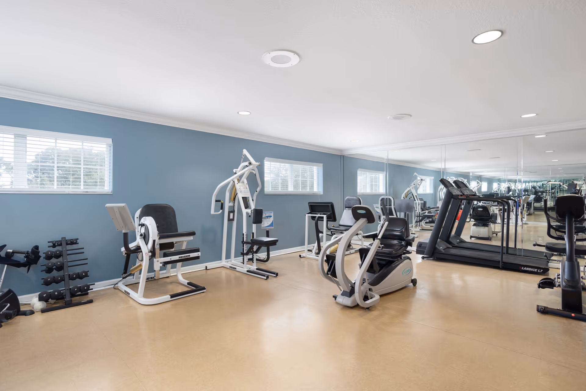 A senior living facility gym room with various exercise equipment including a recumbent bike, weight machines, dumbbells, and treadmills. The room has light blue walls, large mirrors on one side, and windows letting in natural light.