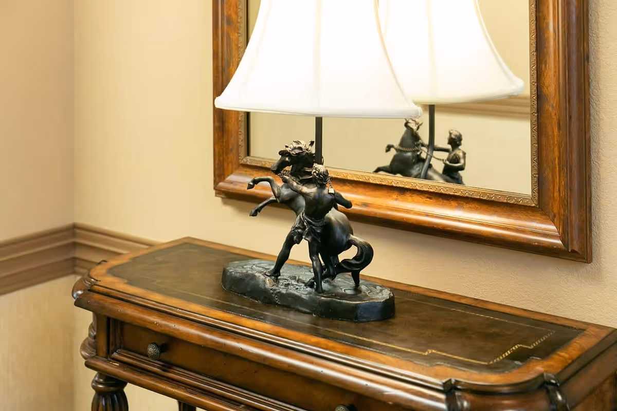 A decorative table lamp with a sculpture of a man and a rearing horse as its base, placed on a wooden console table with a drawer. Behind the lamp is a large wooden-framed mirror reflecting the lamp and sculpture.