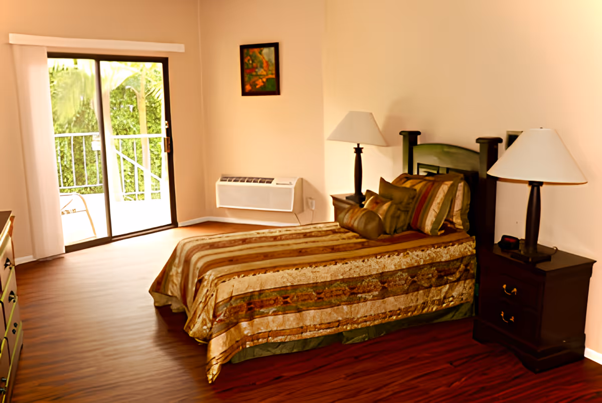 Bedroom with a single bed dressed in striped bedding, two bedside tables with lamps, and sliding glass doors leading to a balcony.