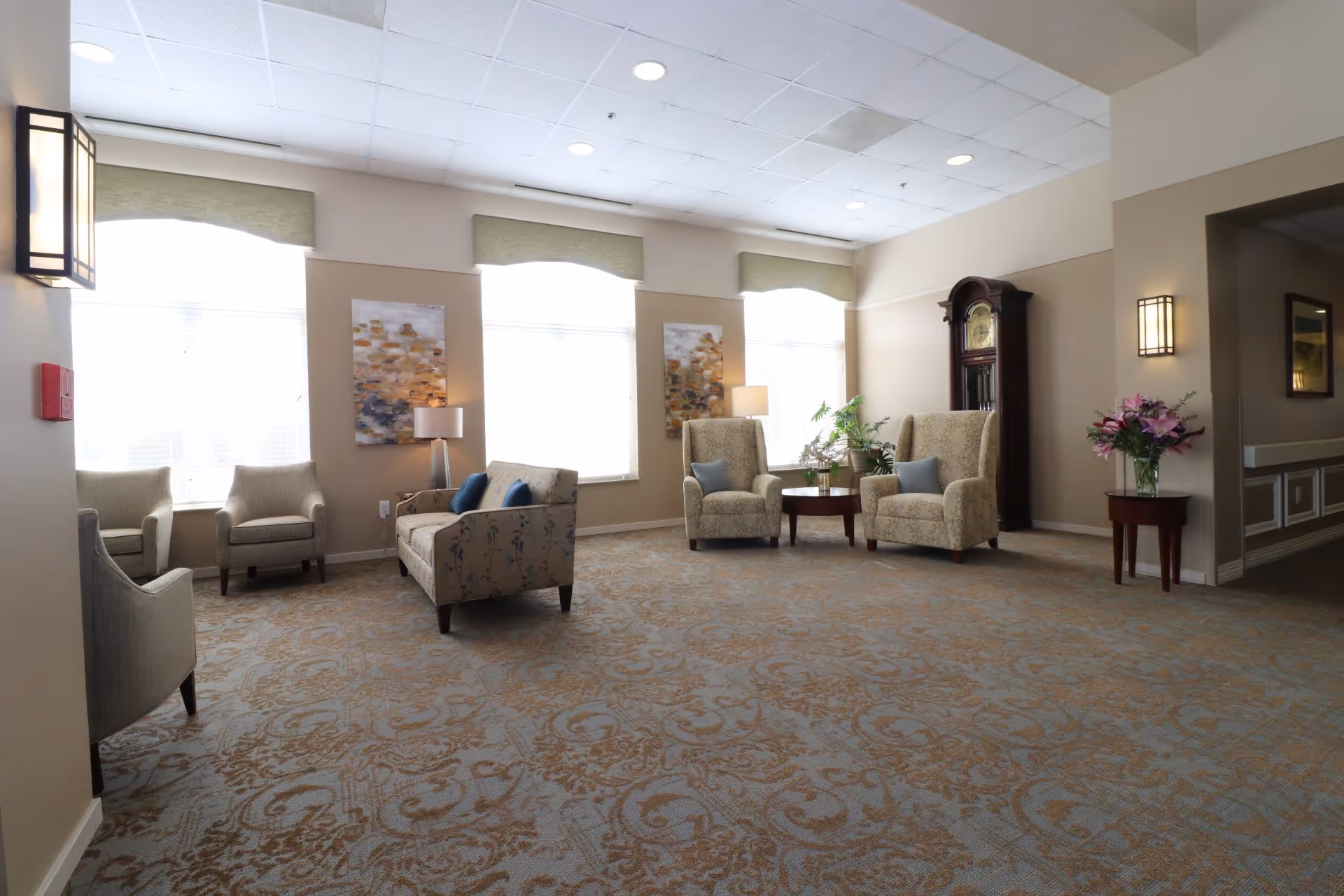 A bright and spacious living room area with patterned carpet, several upholstered armchairs and a loveseat with blue cushions, two large windows with green valances, two abstract paintings on the wall, a tall grandfather clock, a round wooden coffee table with plants, and a small side table with a vase of flowers.