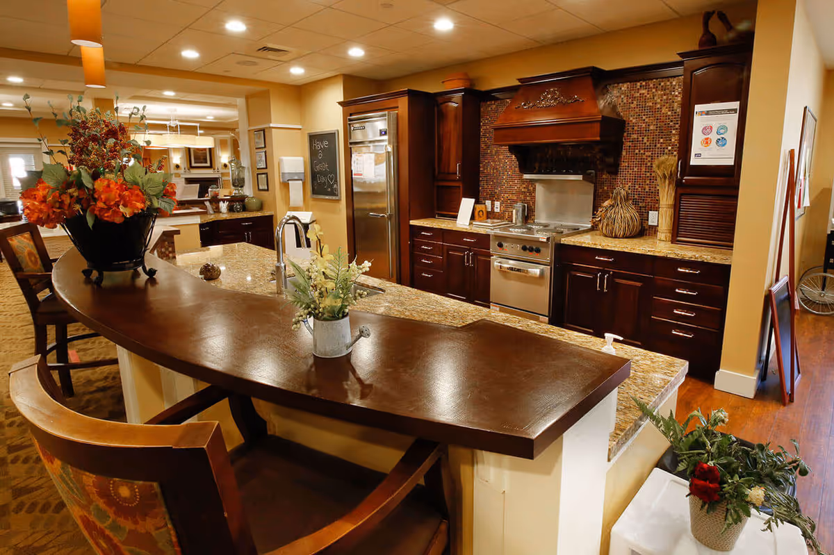 A warm and inviting kitchen area with dark wood cabinets, a stainless steel refrigerator and stove, granite countertops, and a curved wooden breakfast bar with floral arrangements. The space is well-lit with recessed ceiling lights and pendant lights, and there is a chalkboard on the wall with the message 'Have a Great Day'.