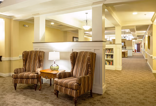 A cozy seating area in a senior living facility with two striped wingback chairs and a wooden side table with a lamp, situated in a well-lit hallway with beige walls, carpeted floor, and built-in bookshelves.