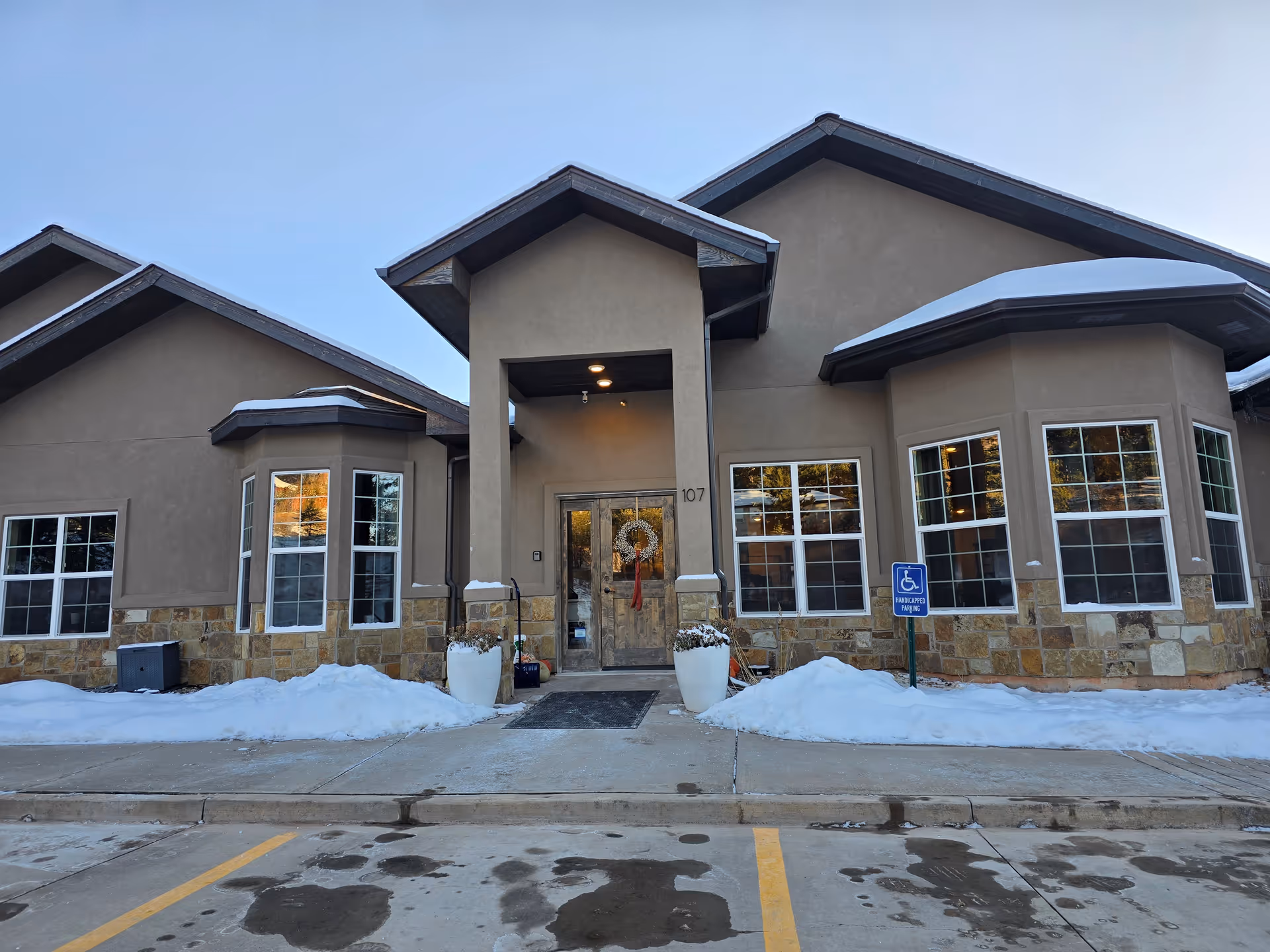 Front entrance of The Aspen building showing a covered entry with double doors, large windows, stone facade, snow along the walkway, and a handicapped parking sign.