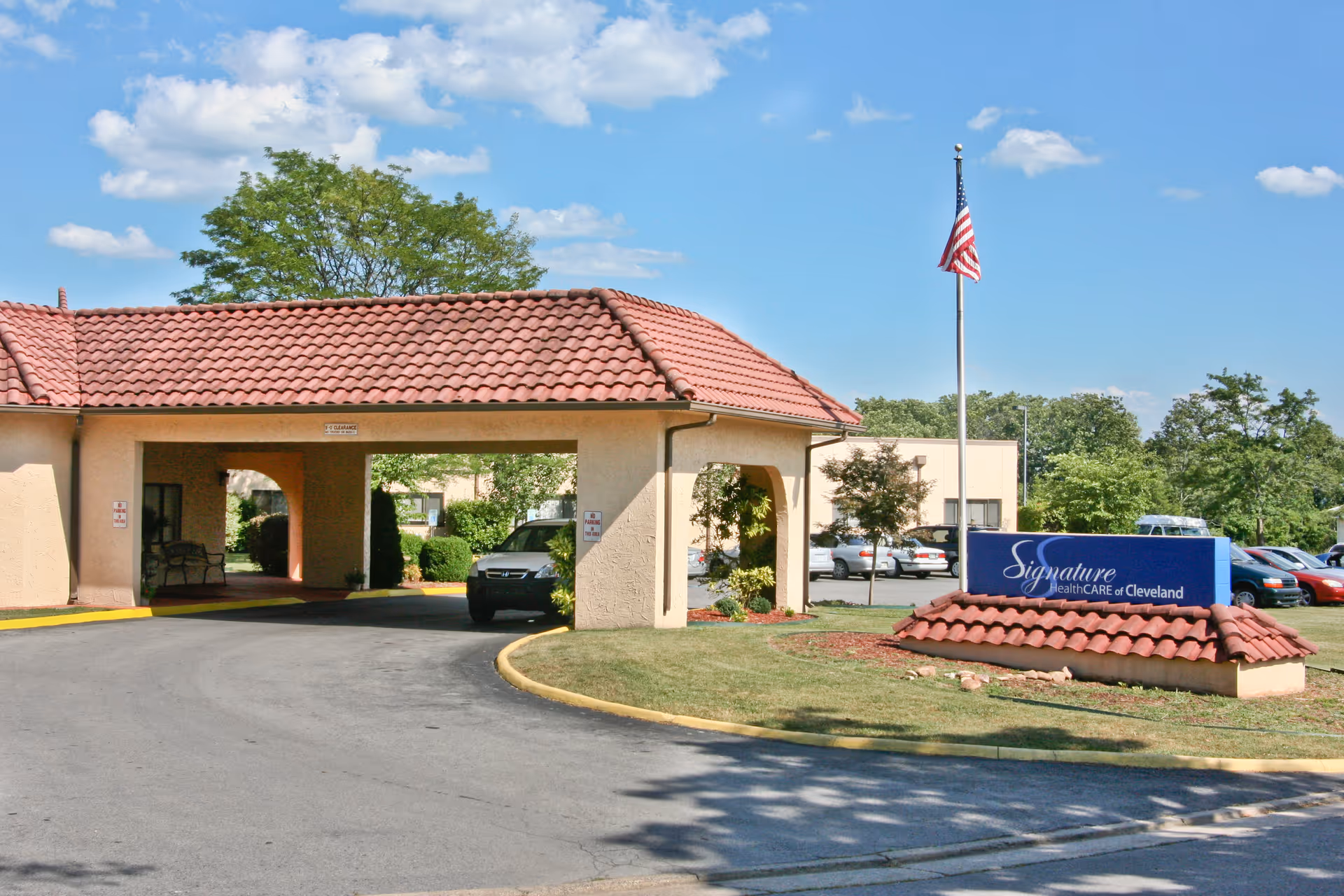 Entrance of Signature HealthCARE of Cleveland facility with a covered driveway, a flagpole with an American flag, a blue sign displaying the facility name, and a parking lot with several cars in the background under a partly cloudy sky.