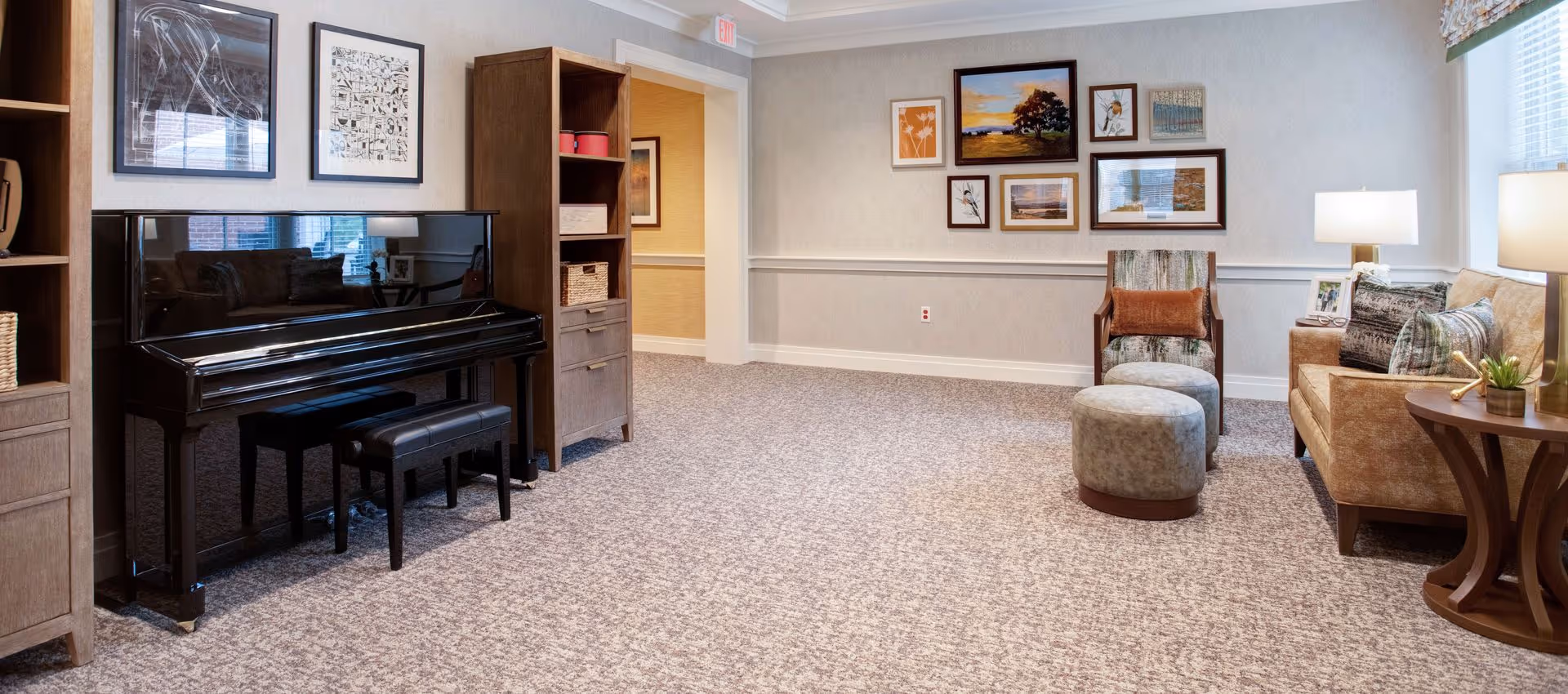 Community lounge with an upright piano, chairs, ottomans, and framed artwork on the walls.