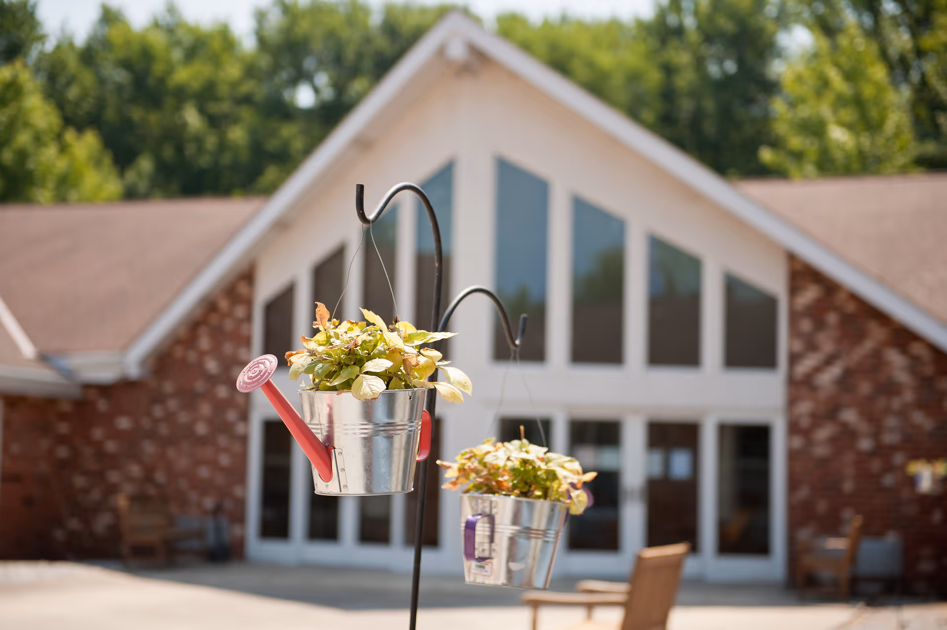 Two small metal buckets with plants hanging from black metal hooks in front of a brick building with large triangular windows and a brown roof, surrounded by greenery.