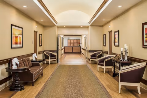 A well-lit hallway in a senior living facility with a high arched ceiling, beige walls, and wooden flooring. The hallway is furnished with several chairs and small tables on both sides, decorated with framed artwork and candle holders. At the end of the hallway, there is a reception desk area.