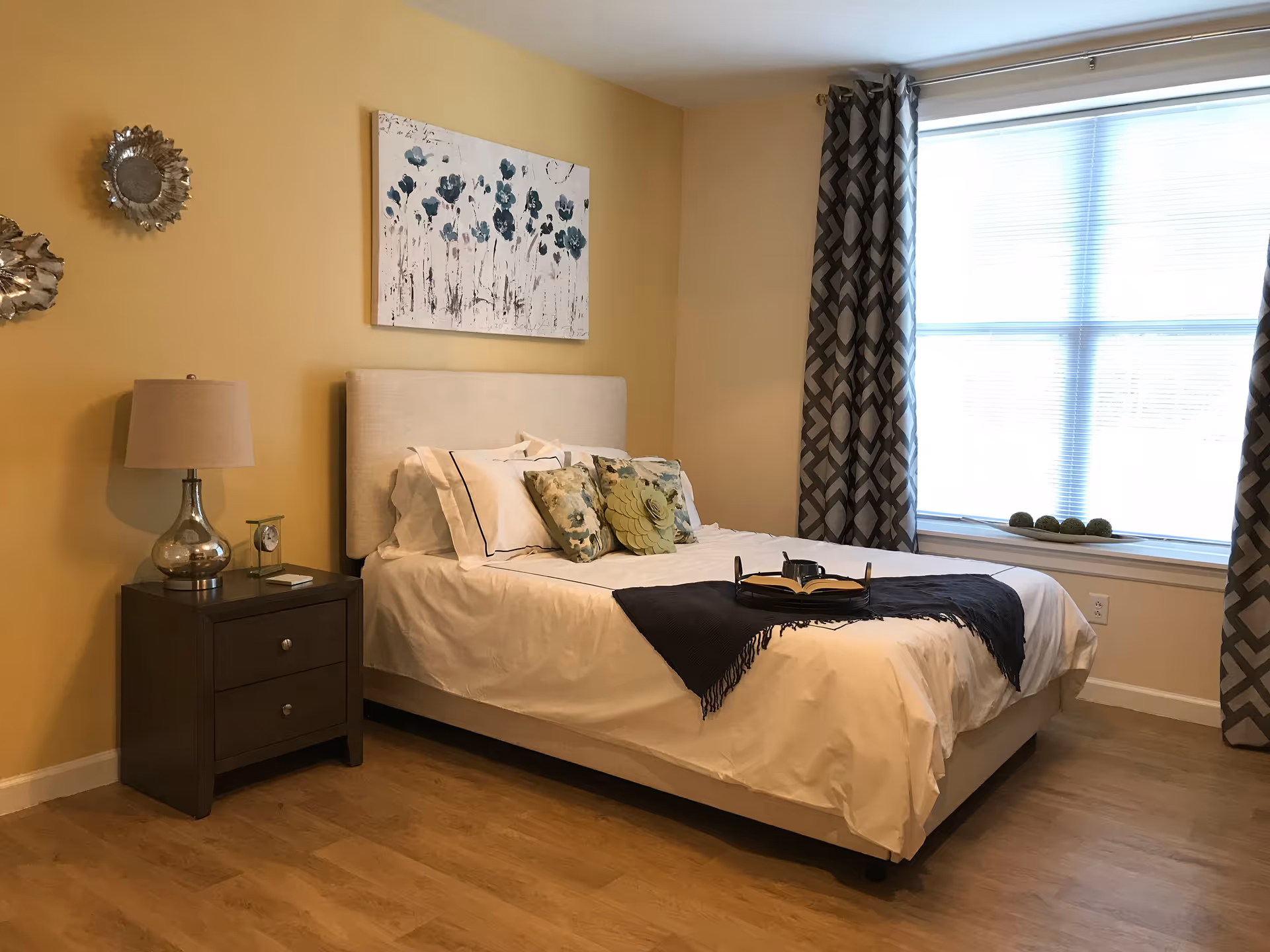 A cozy bedroom with a neatly made bed featuring white linens, decorative pillows, and a dark throw blanket. There is a wooden nightstand with a lamp and a small clock on it. The walls are painted yellow, and there is a floral painting above the bed. A large window with patterned curtains lets in natural light.