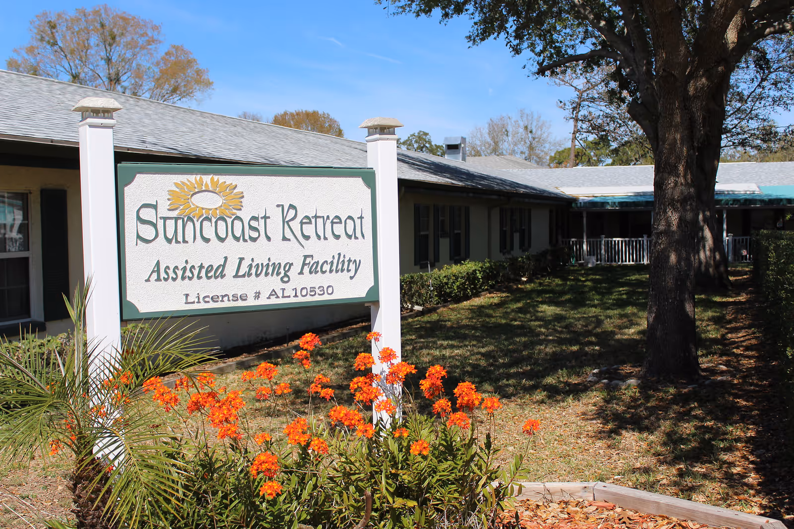 Outdoor view of Suncoast Retreat Assisted Living Facility sign with orange flowers in front and a single-story building with windows and a tree in the background under a clear blue sky.