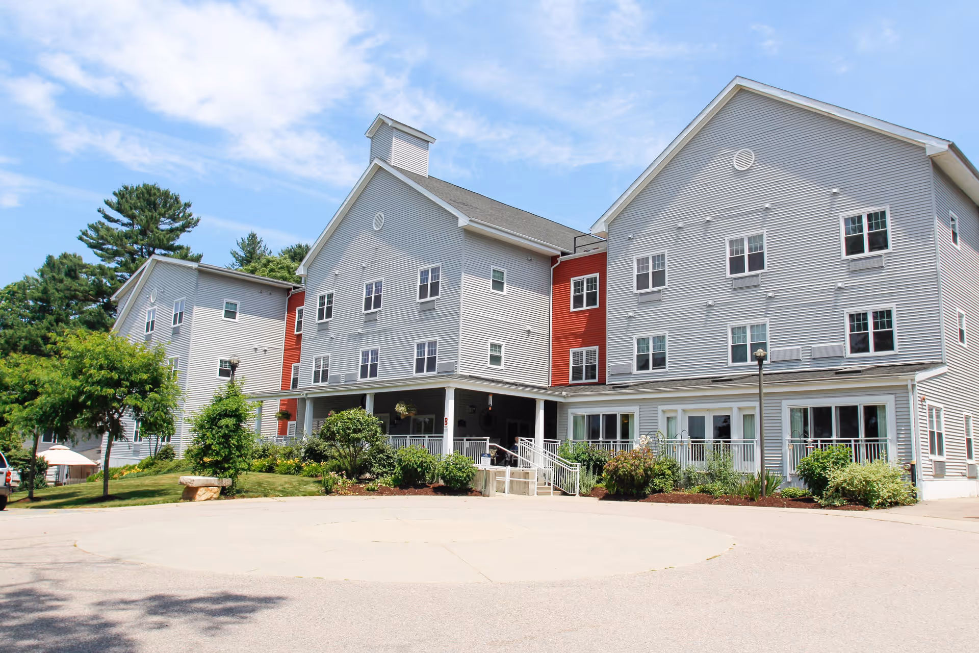Exterior view of a large, three-story senior living facility building with gray siding and red accents. The building has multiple windows, a covered entrance with a ramp, and surrounding greenery including bushes and trees under a partly cloudy sky.