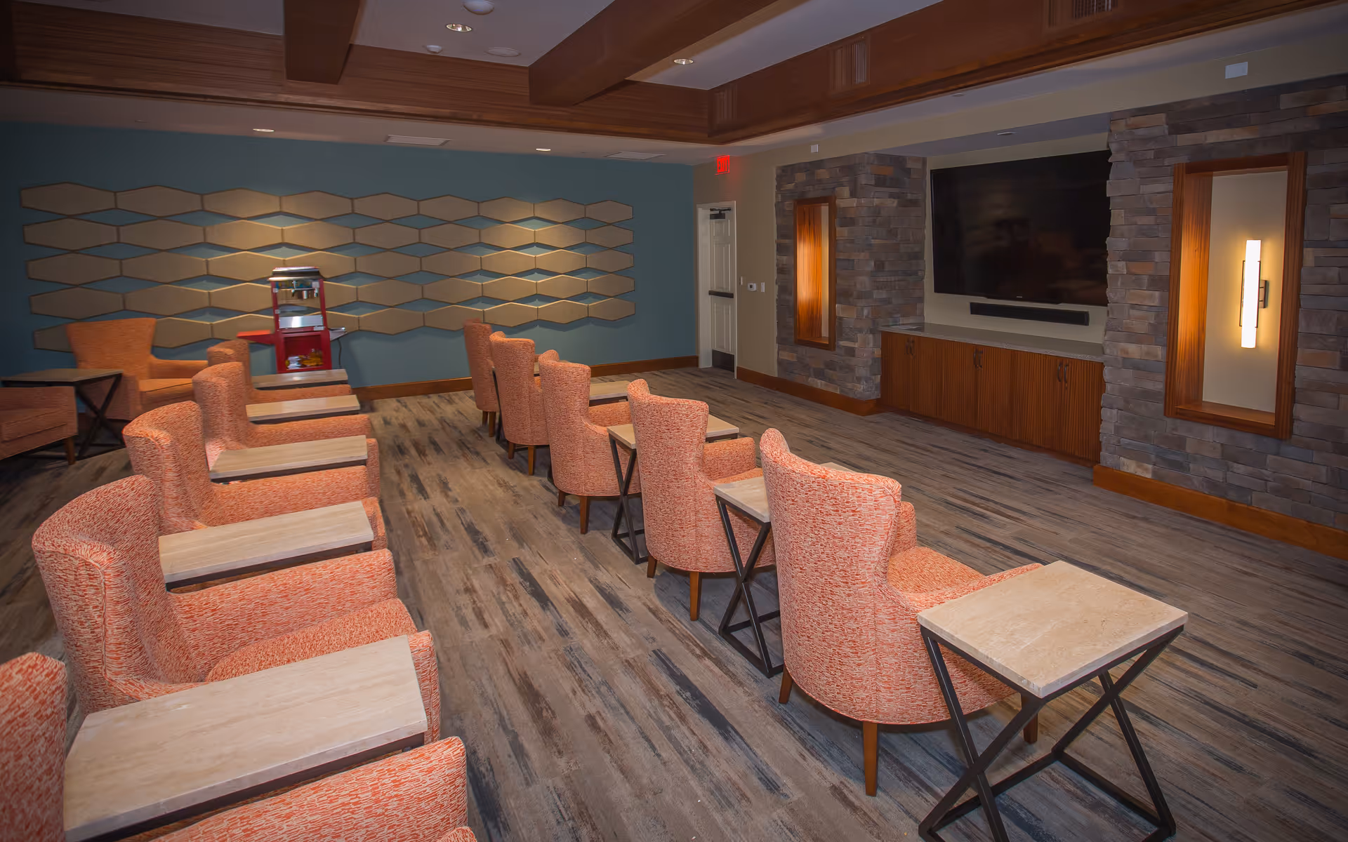 Rows of orange upholstered chairs with individual side tables facing a large wall-mounted TV in a communal media/lounge room.