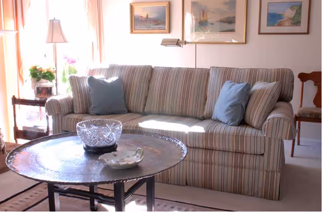 Sunlit living room with a striped sofa and blue pillows, a round coffee table, lamp, and framed paintings on the wall.
