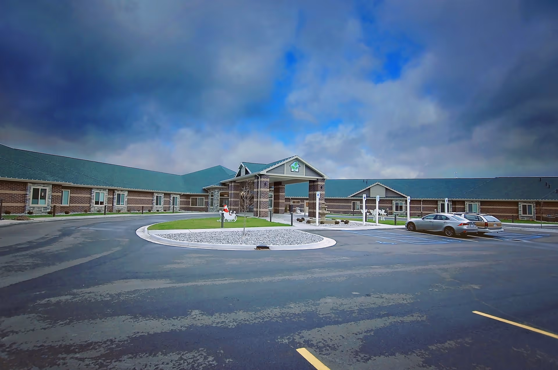 Exterior view of a single-story senior living facility with a green roof and brick walls under a cloudy sky. There is a circular driveway with a small landscaped island in the center, and two cars are parked in designated parking spaces near the entrance.