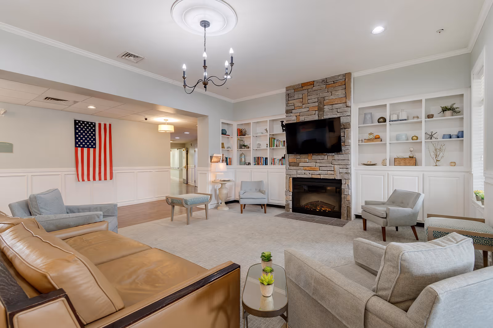 A cozy living room with a stone fireplace and a mounted flat-screen TV above it. The room features built-in white shelves filled with decorative items and books. There are several comfortable chairs and a tan leather sofa arranged around a small glass coffee table with potted plants. An American flag hangs on the wall in the background, and a chandelier light fixture hangs from the ceiling.