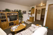 A cozy living room in a senior living facility featuring a floral-patterned sofa, a wooden coffee table with various items on it, a wooden entertainment center with a television and decorative items, and a small kitchenette area in the background with cabinets and a refrigerator.