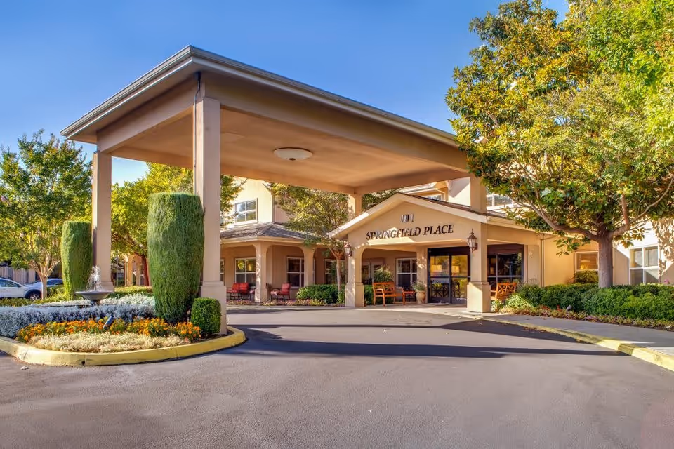 Entrance of Springfield Place senior living facility with a covered drop-off area, landscaped garden with flowers and shrubs, and benches near the entrance under a clear blue sky.