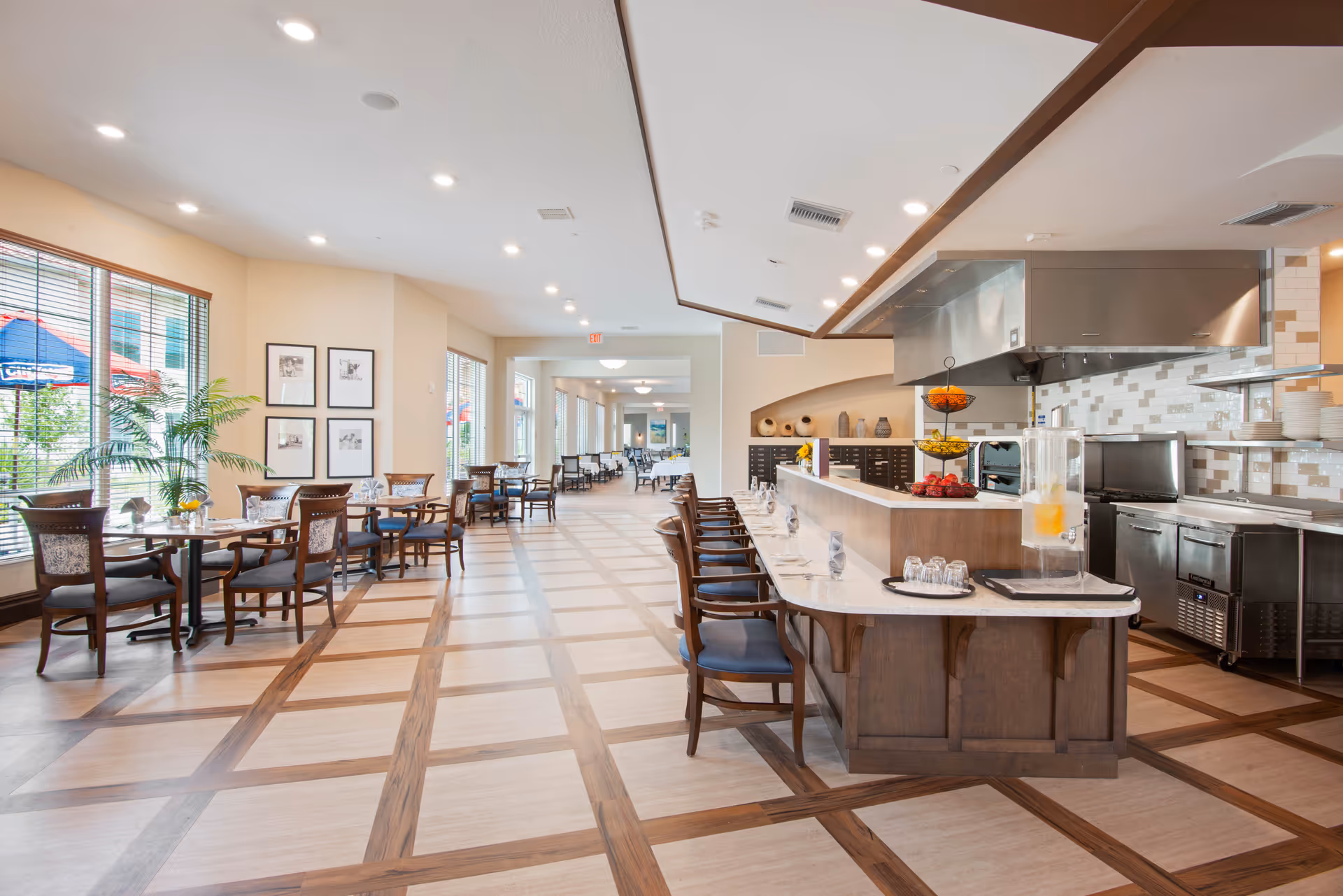 A spacious dining area in a senior living facility featuring a long counter with bar stools on one side and several small dining tables with chairs on the other. The room has large windows with blinds, framed pictures on the wall, and a modern kitchen area with stainless steel appliances and a tiled backsplash. The floor has a checkered wood pattern, and the space is well-lit with recessed ceiling lights.