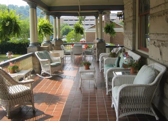 Covered front porch with white wicker chairs and tables, potted plants and hanging ferns on a tiled floor overlooking a lawn.