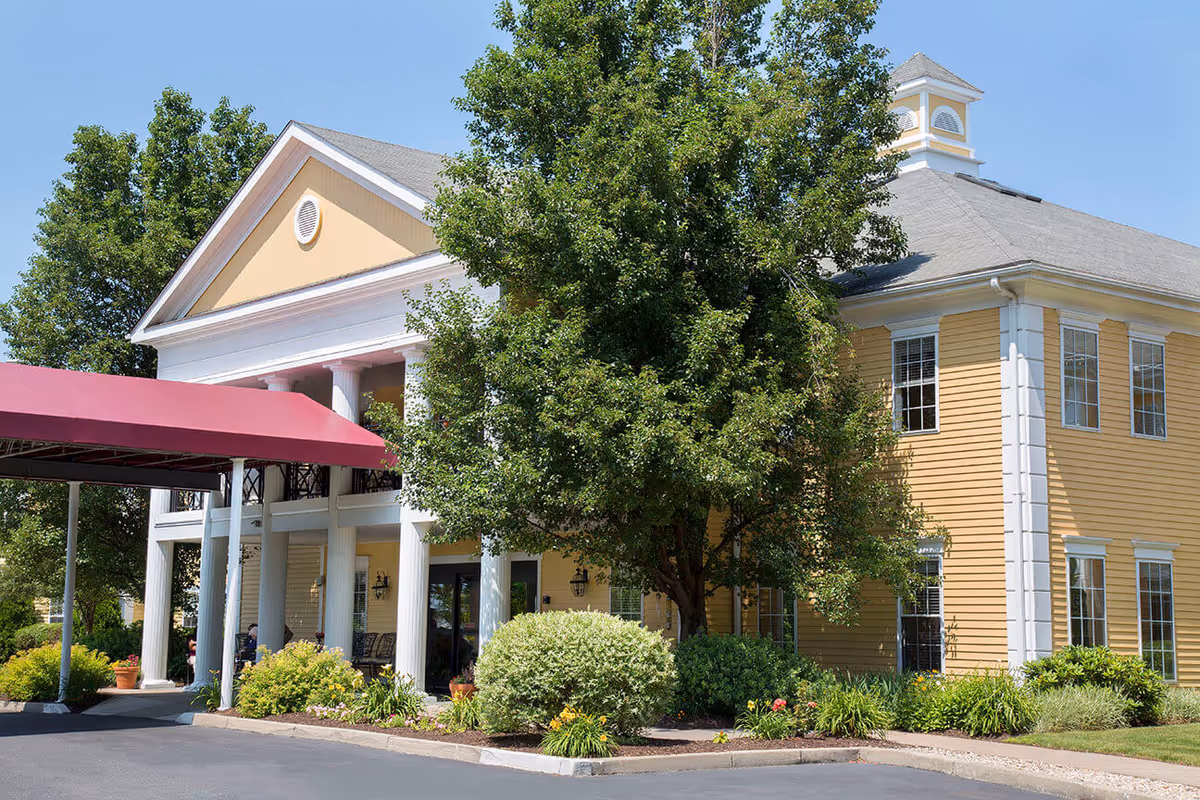Front exterior of a yellow senior living building with a covered entrance, white columns, and surrounding landscaping.