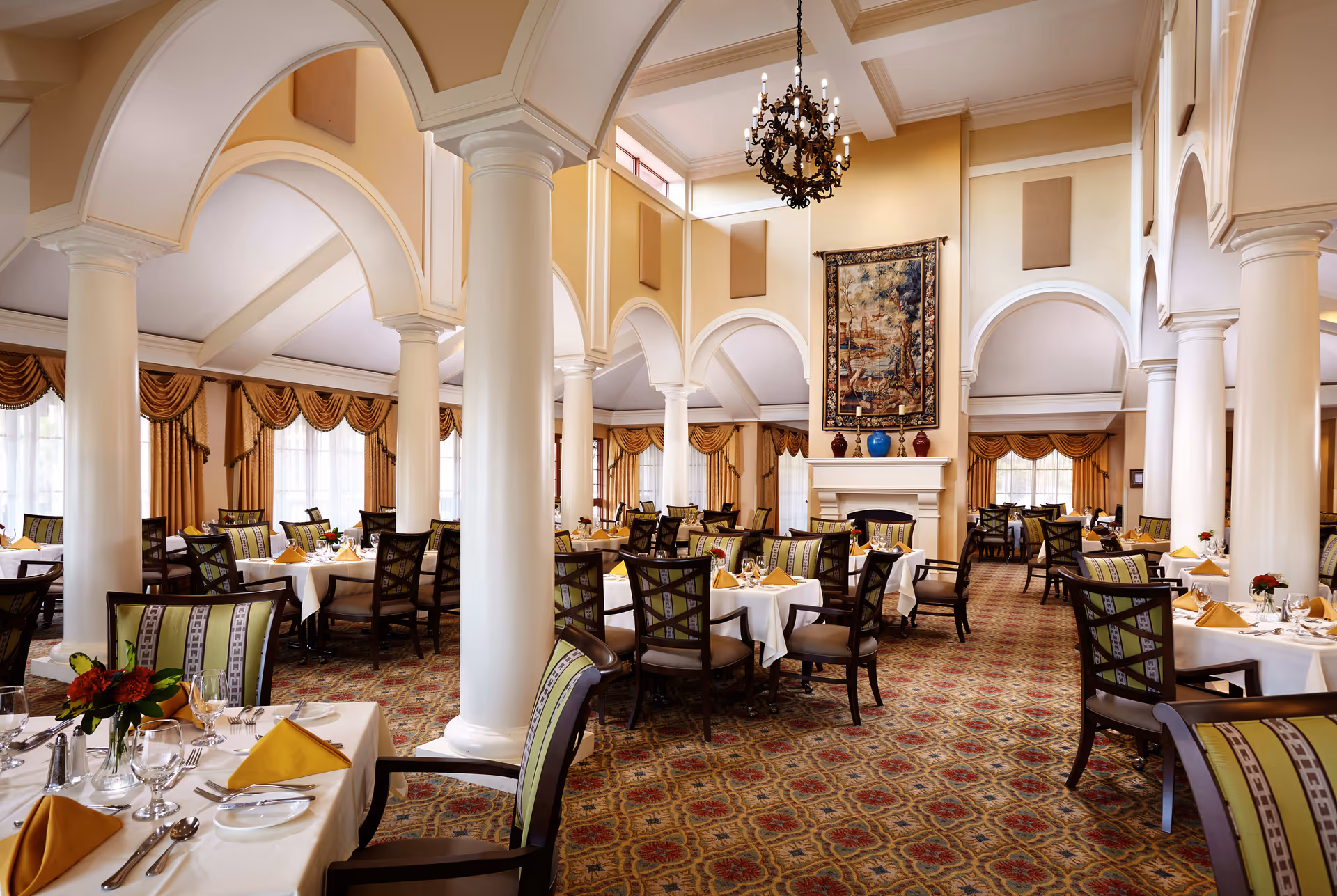 Elegant dining room with high vaulted ceilings, white columns, and arched architectural details. Tables are set with white tablecloths, yellow napkins, glassware, and silverware. The room features patterned carpet, large windows with gold drapes, a chandelier, and a decorative tapestry above a fireplace.