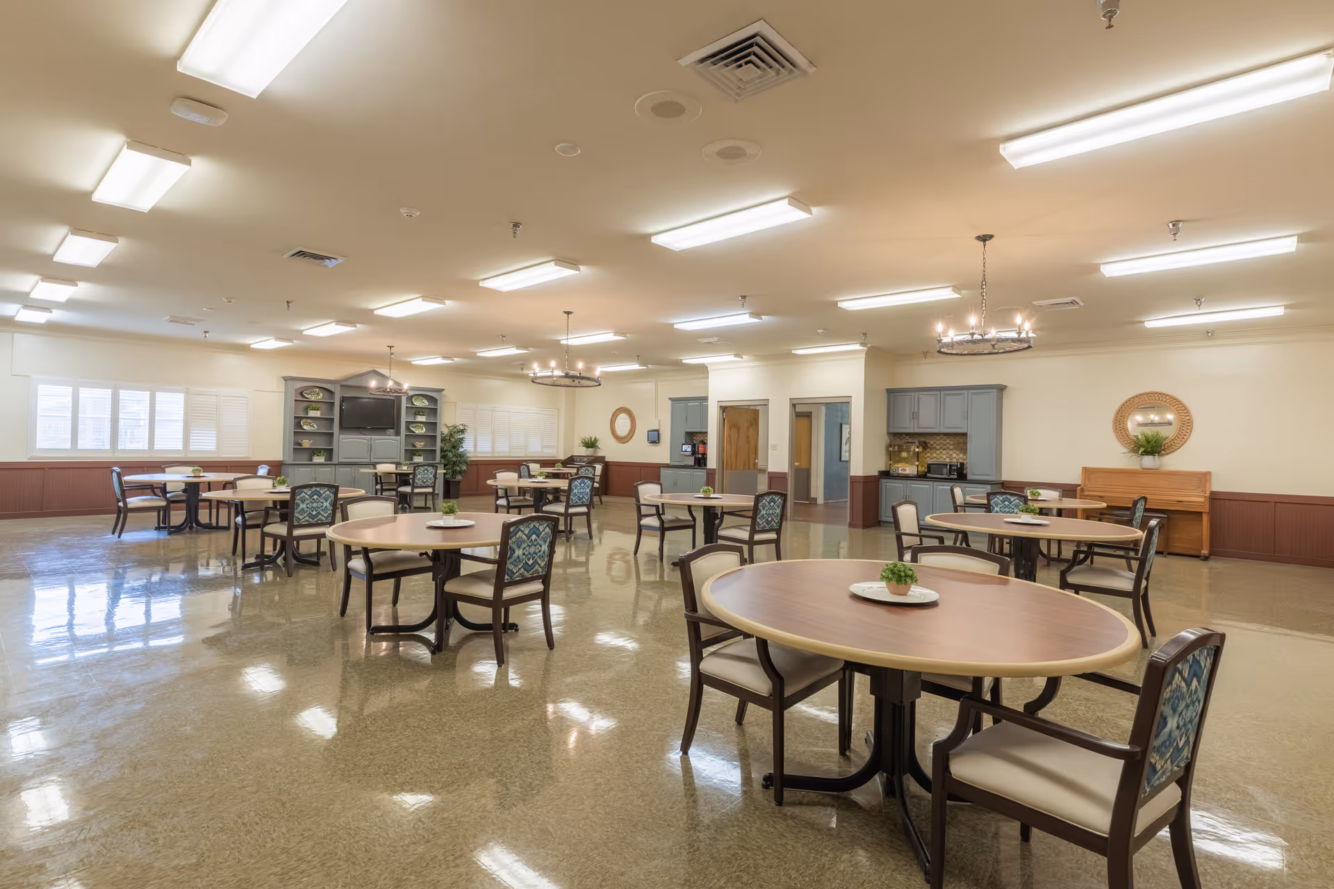 A spacious dining room with multiple round tables and chairs arranged neatly. Each table has a small decorative plant centerpiece. The room has bright overhead lighting, large windows with white shutters, a cabinet with a television, and a small kitchenette area with blue cabinets. The walls are painted cream with a brown wainscoting, and there are chandeliers hanging from the ceiling.