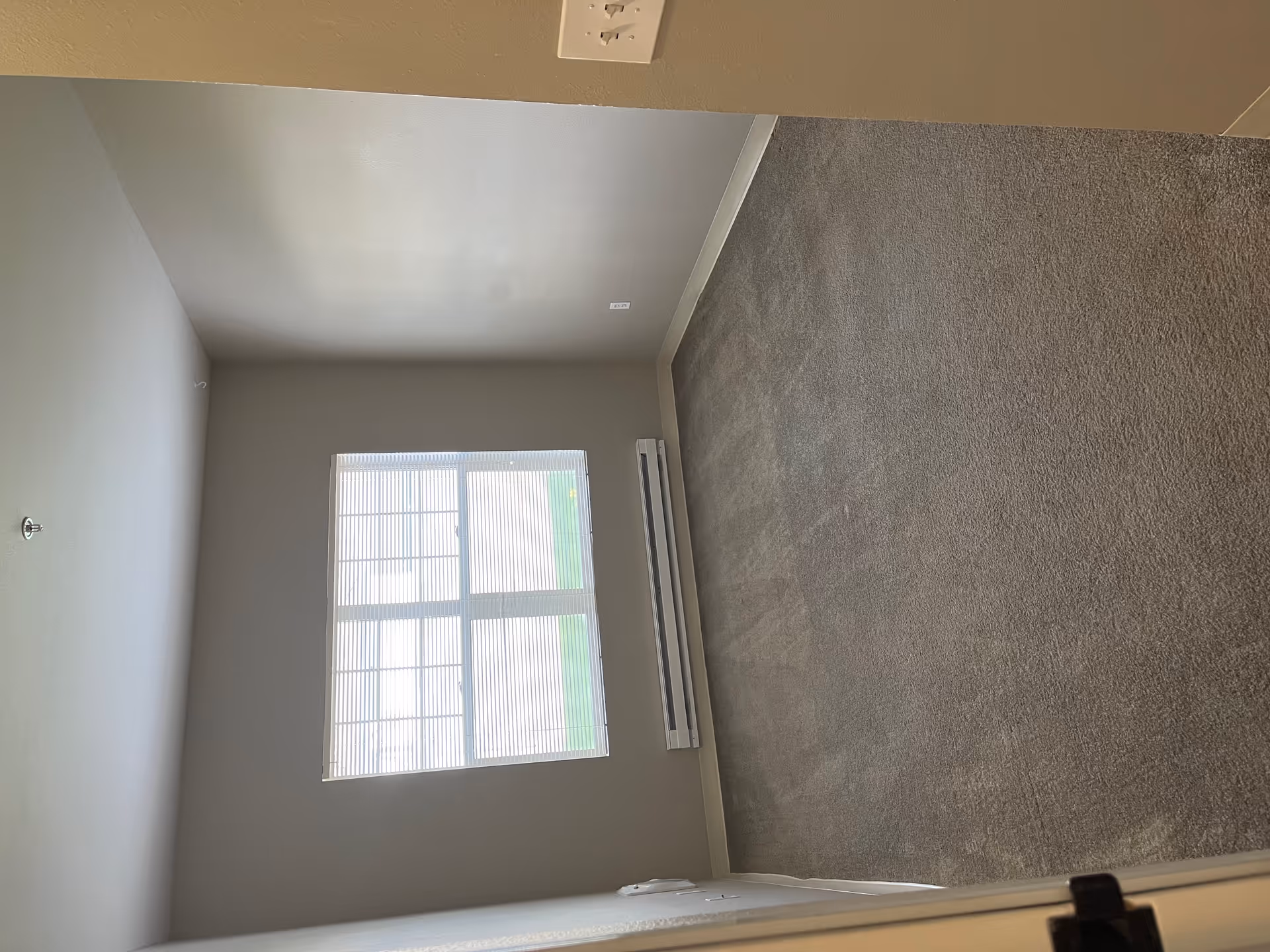 Empty carpeted bedroom with a window with closed blinds, a baseboard heater, and beige walls.