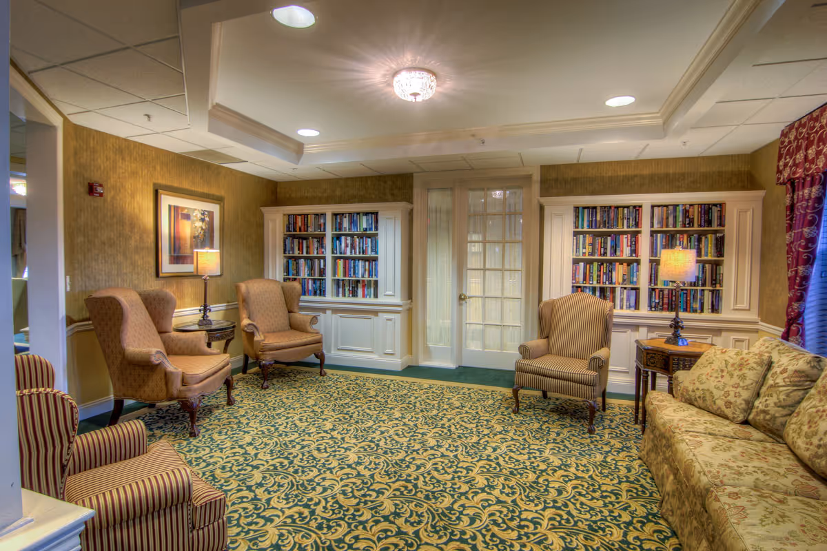 A cozy living room area with patterned carpet, two built-in bookshelves filled with books, a floral sofa, three upholstered armchairs, two side tables with lamps, and a ceiling light fixture. The walls are painted in warm tones and there are French doors in the background.