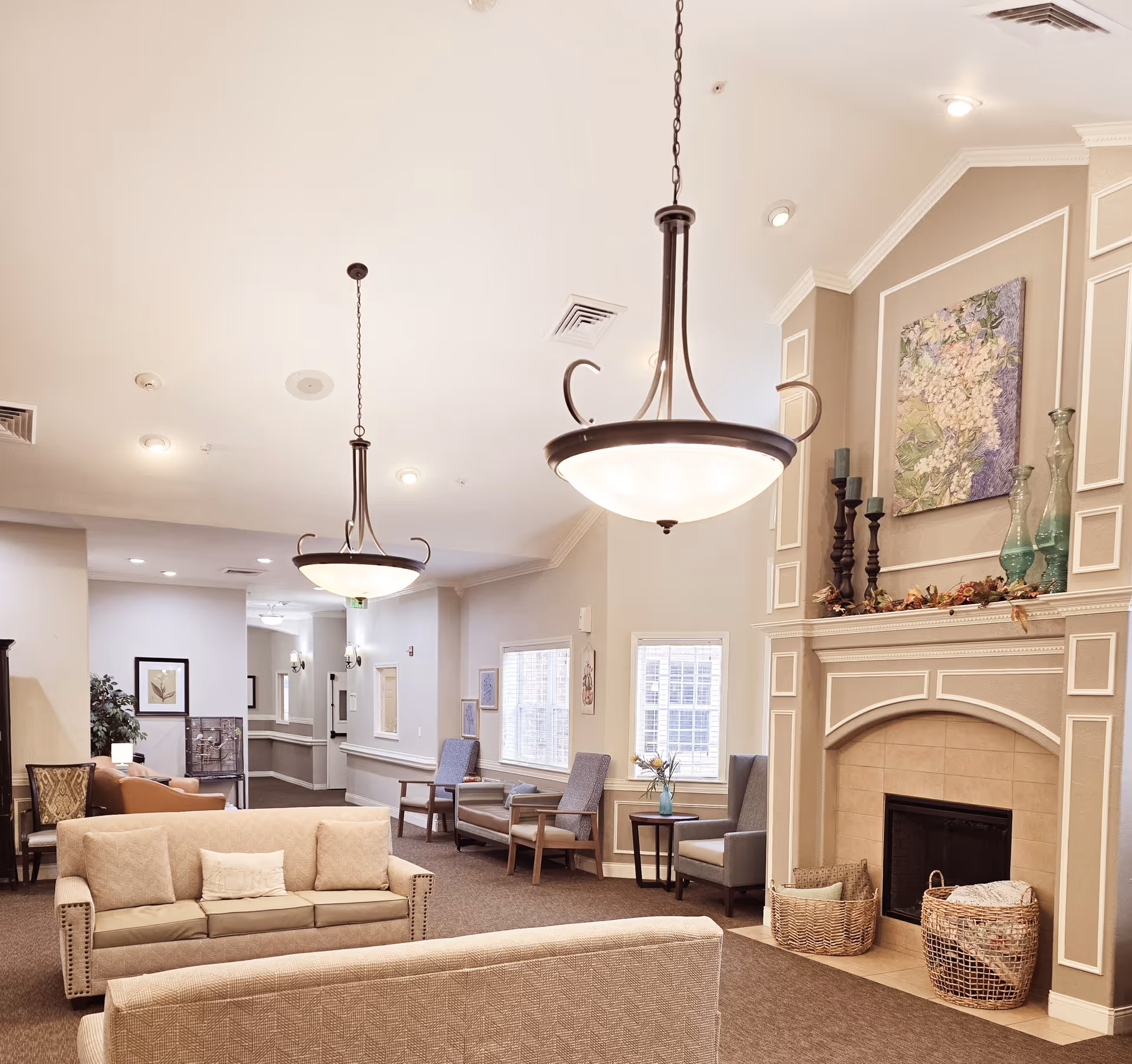 A cozy and well-lit living room area in a senior care facility featuring two beige sofas, several armchairs along the wall, a fireplace with decorative baskets and vases on the mantel, and two large hanging light fixtures from the ceiling. The walls are painted in neutral tones with framed artwork and windows letting in natural light.