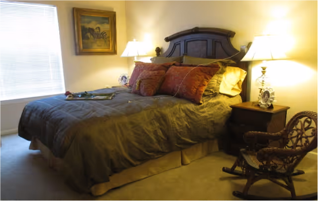A furnished bedroom featuring a large bed with decorative pillows, two bedside tables with lamps, a woven rocking chair, and a window with blinds.