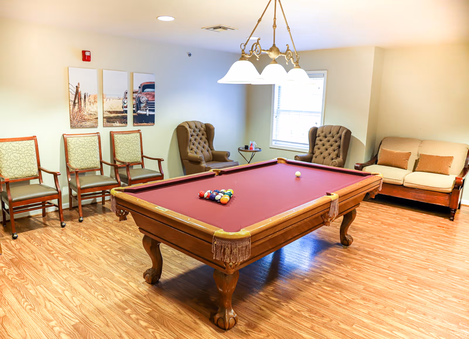 A bright recreation room centered on a red-felt pool table with surrounding chairs, armchairs, and a sofa under overhead lights.