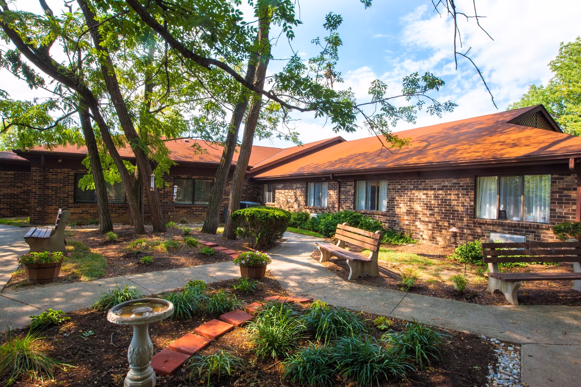 Brick single-story care center building with a shaded courtyard featuring benches, trees, a birdbath, and walkways.