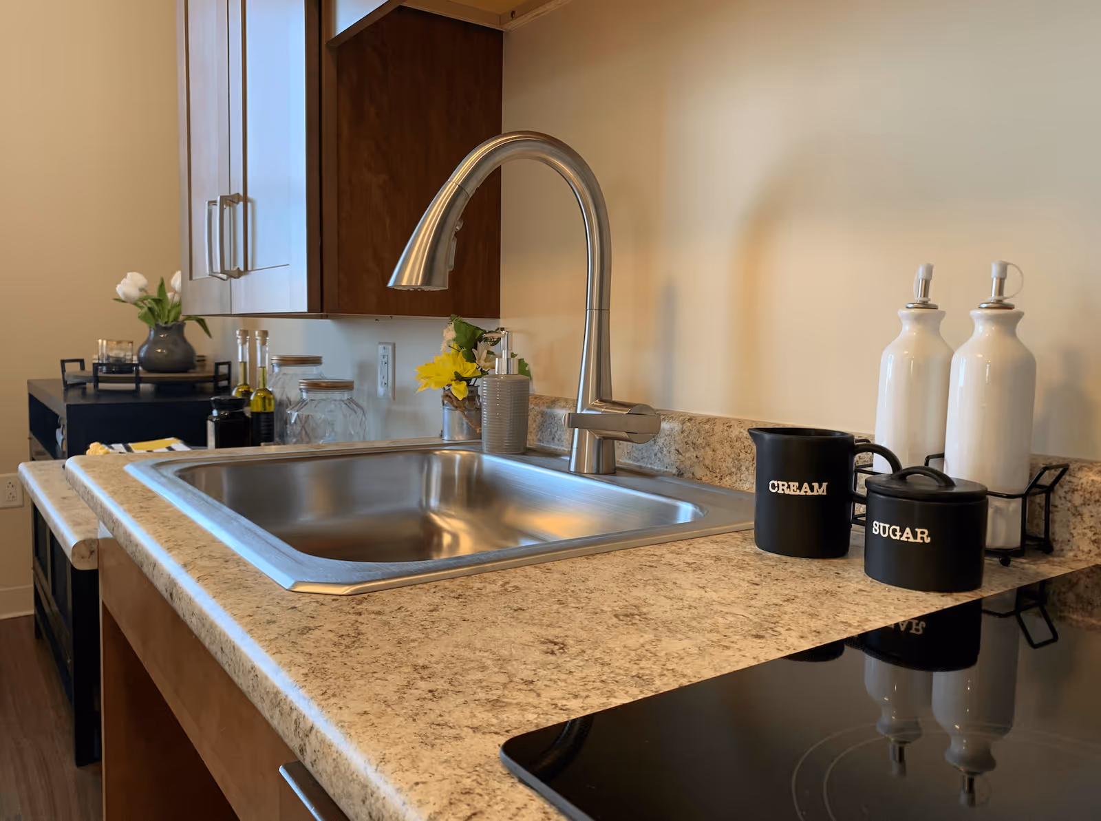 A clean kitchen countertop with a stainless steel sink and modern faucet. On the right side of the sink are black containers labeled 'CREAM' and 'SUGAR' along with two white bottles with dispensers. In the background, there are wooden cabinets, a small vase with flowers, and some kitchen items on a black cart.