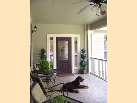 Covered front porch featuring a purple front door, seating, potted plants, and a dog lying on the tiled floor.