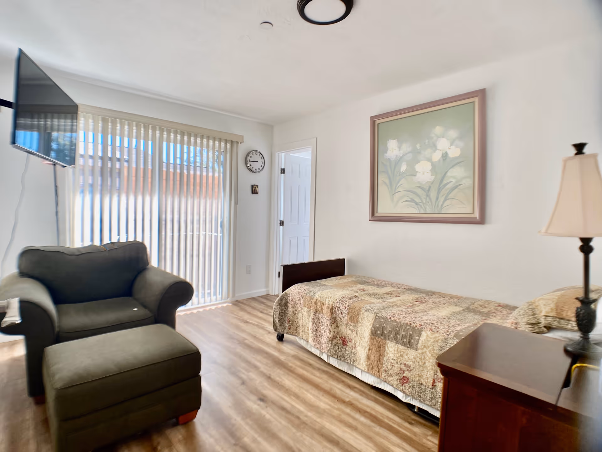 A cozy room with a single bed covered in a patchwork quilt, a wooden nightstand with a lamp, a green armchair with a matching ottoman, a wall-mounted TV, a clock on the wall, and vertical blinds covering a sliding glass door letting in natural light.