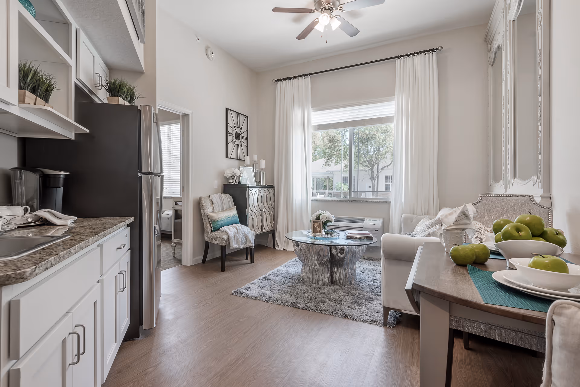 A bright and cozy living space featuring a small kitchen area with white cabinets and granite countertops on the left, a stainless steel refrigerator, and a coffee maker. The room opens into a living room with a large window covered by white curtains, a ceiling fan, a gray area rug, a round wooden coffee table, a white sofa, and a decorative chair with a throw blanket. A dining table with green apples and white plates is visible on the right side. The walls are light-colored, and the floor is wood.