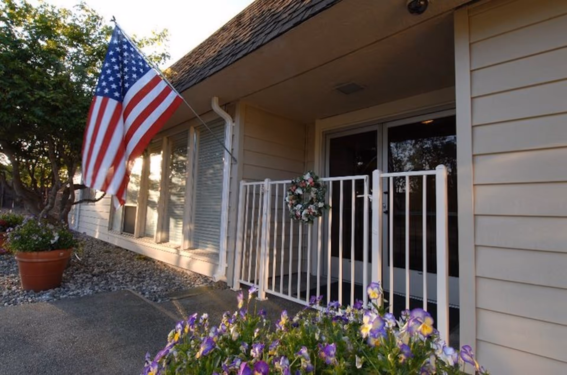 Entrance to a building with beige siding, a white gate decorated with a floral wreath, an American flag mounted on the wall, and purple flowers in the foreground.