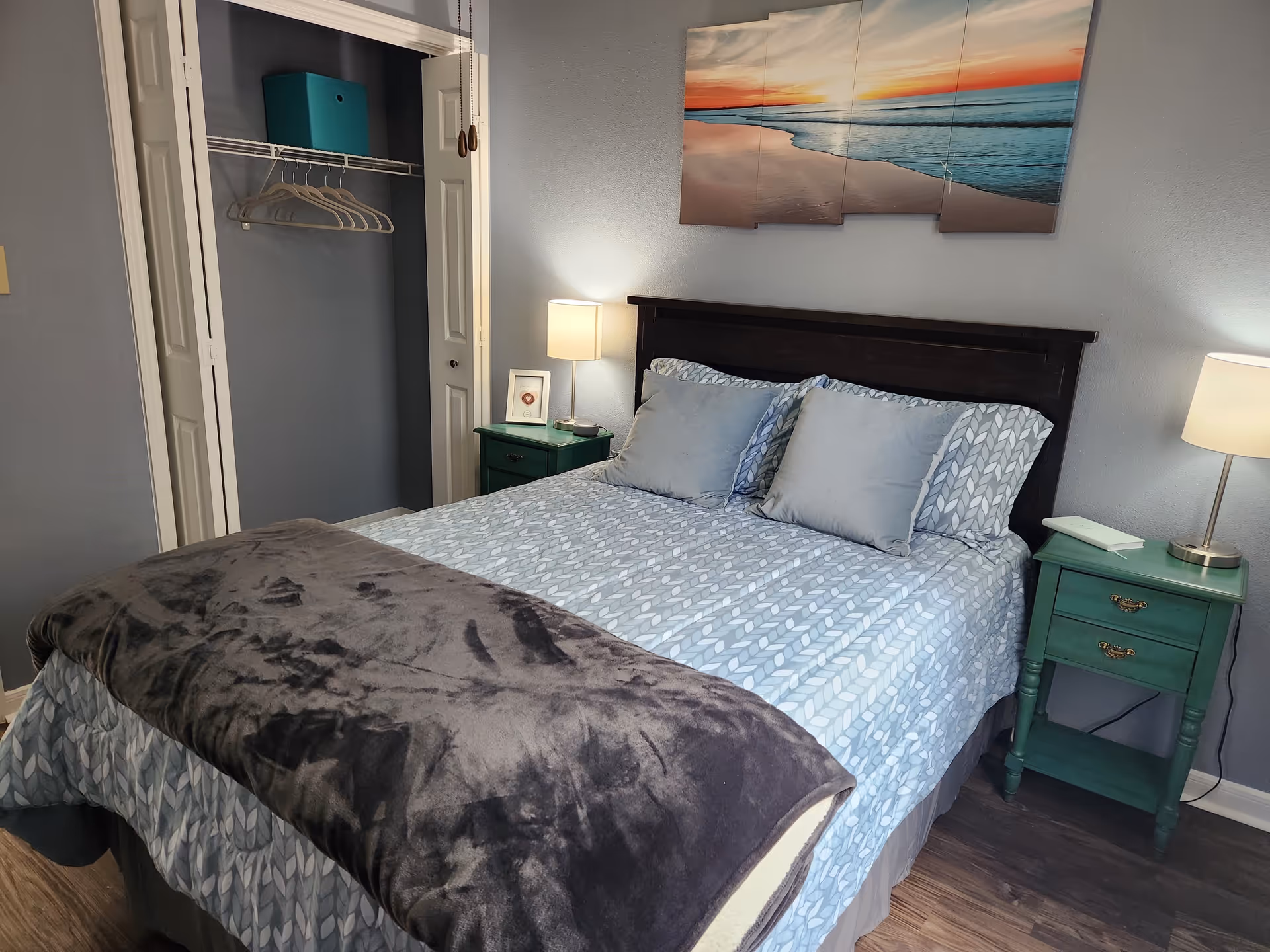 A neatly made bed with a patterned blue and white bedspread and two gray pillows in a bedroom. There is a dark wooden headboard, two green nightstands with lamps on each side of the bed, and a multi-panel wall art depicting a beach sunset above the headboard. An open closet with hangers and a teal storage box is visible to the left.
