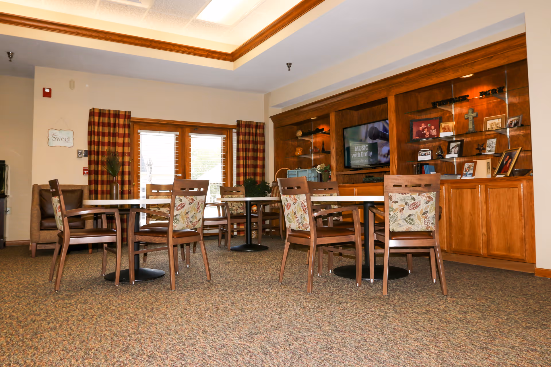 A senior living common area with round tables and chairs in front of wooden built-in shelving and a television.