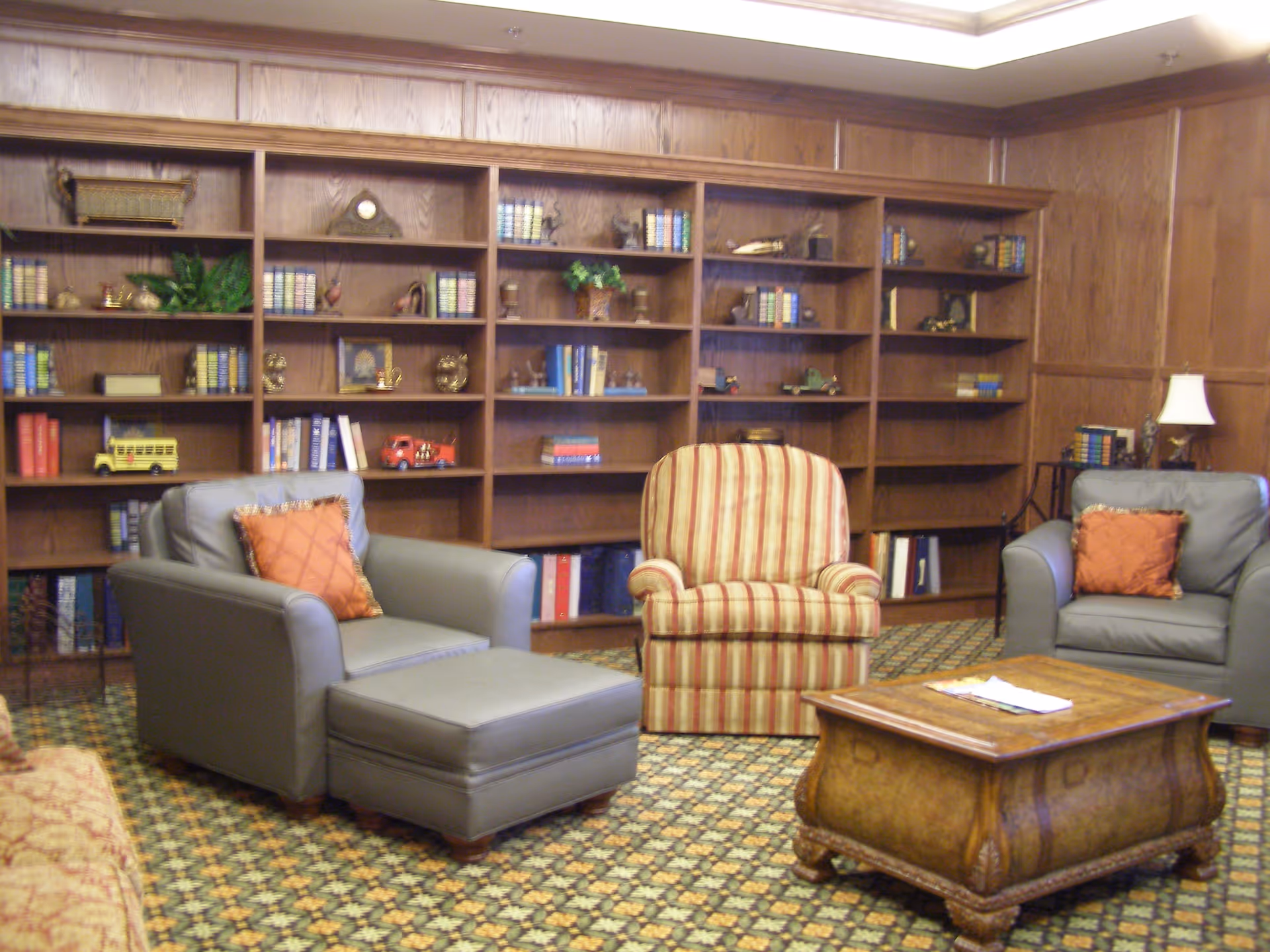 A cozy common room with upholstered armchairs, a striped accent chair, ottoman, wooden coffee table and built-in bookshelves.