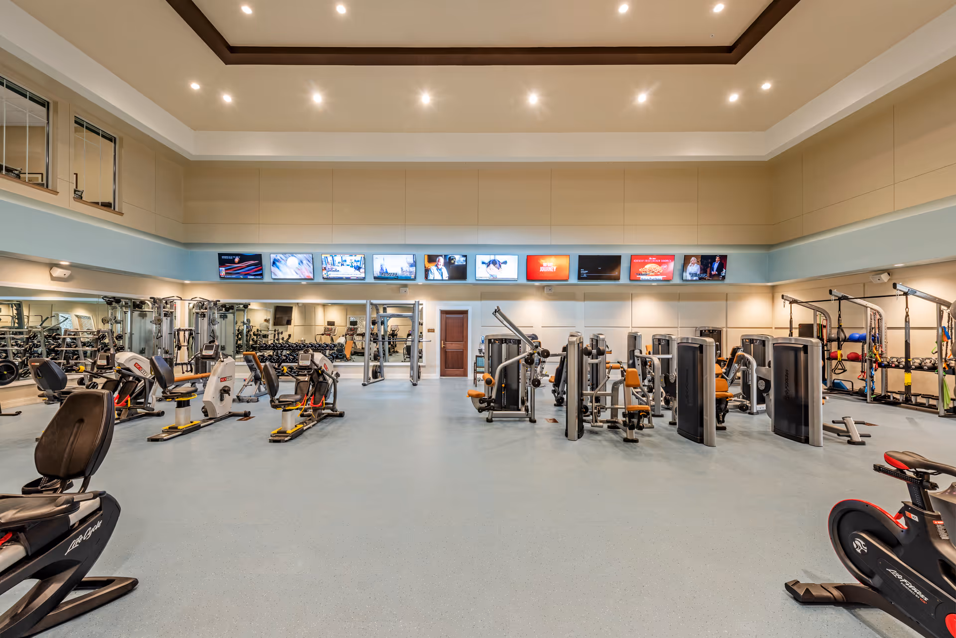 A spacious fitness center with various exercise machines including stationary bikes, weight machines, and resistance training equipment. The room has high ceilings with recessed lighting, a row of televisions mounted on the wall, and large mirrors along one side.