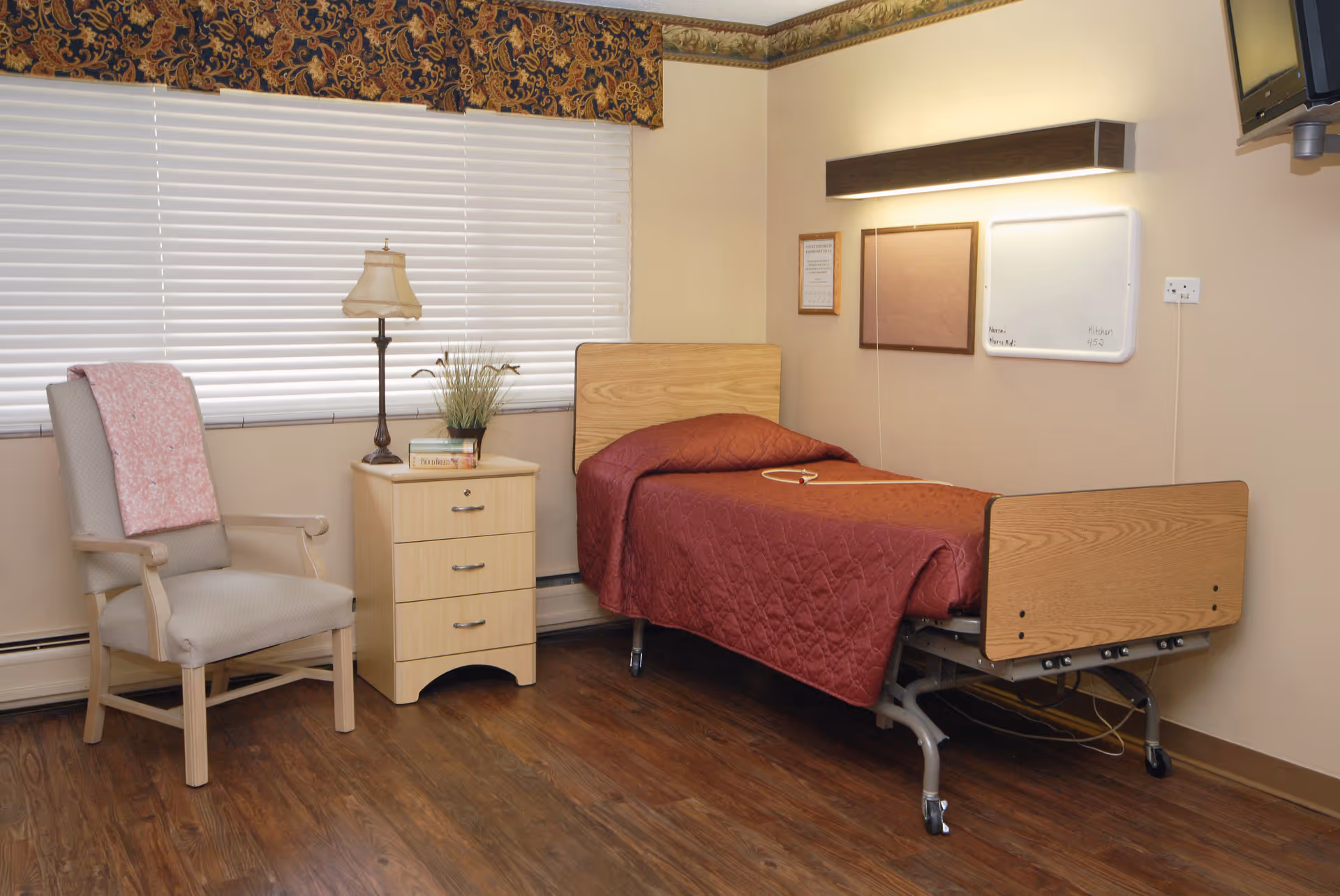 A simple senior living bedroom with a single hospital-style bed covered in a red quilt, a beige armchair with a pink blanket draped over it, a small wooden nightstand with a lamp, books, and a plant, a window with closed blinds and a decorative valance, and a wall-mounted TV in the corner.