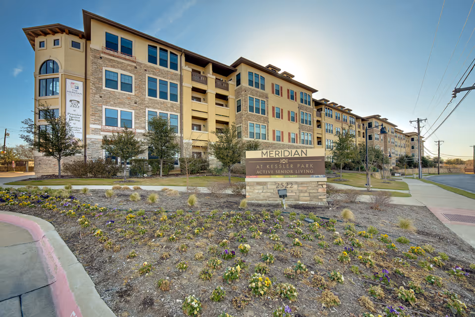Exterior view of a multi-story senior living facility building named Meridian at Kessler Park with a landscaped flower bed and a sign in front displaying the facility name and address 2522. The building has multiple windows, balconies, and a mix of stone and stucco facade under a clear blue sky.