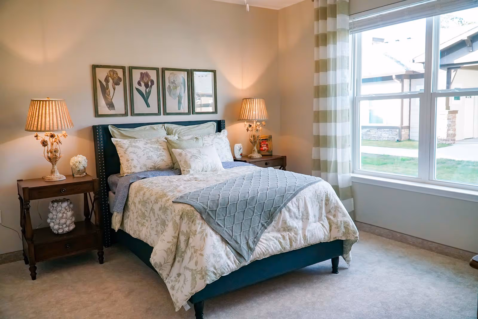 A cozy bedroom with a neatly made bed featuring floral patterned bedding and multiple pillows. There are two wooden nightstands on either side of the bed, each with a lamp and decorative items. Above the bed, there are four framed botanical prints. A large window with striped curtains lets in natural light and shows a view of the outside buildings and lawn.