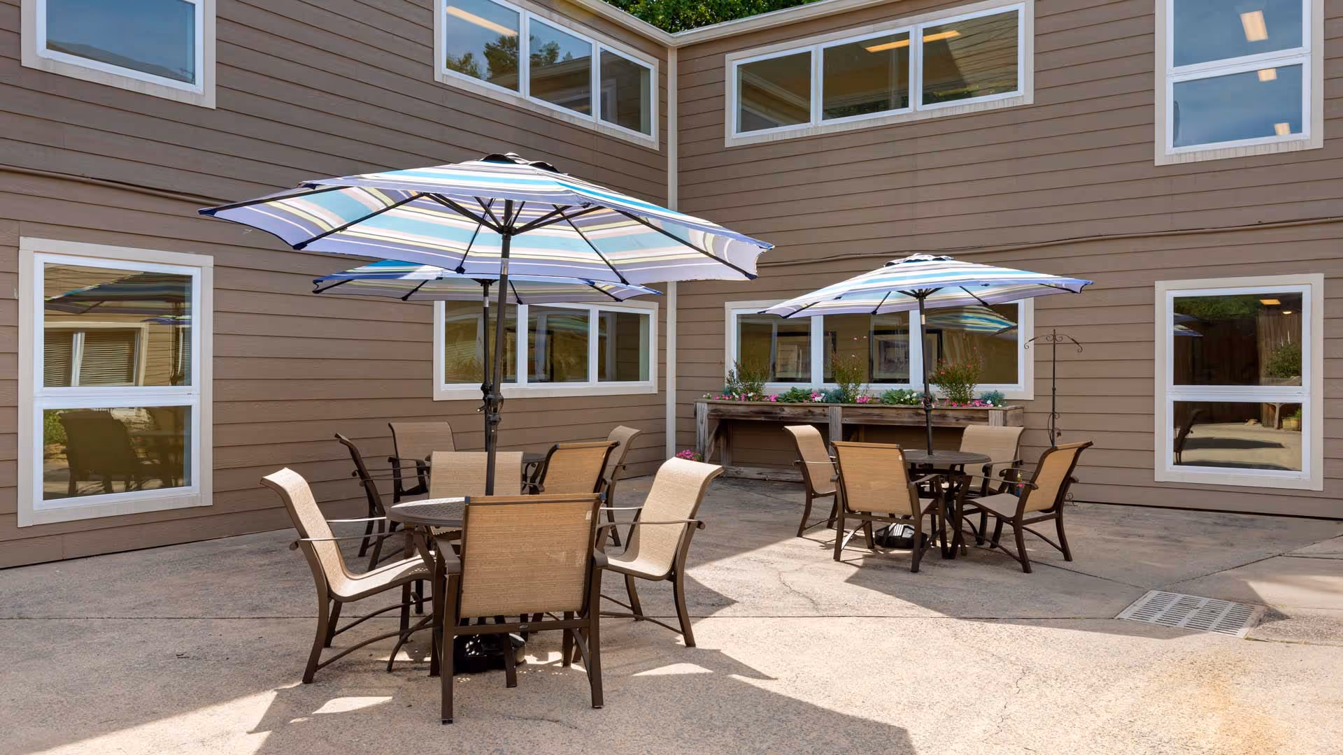 Outdoor patio area with two round tables, each surrounded by four chairs and shaded by large striped umbrellas. The patio is enclosed by a building with multiple windows and a planter box with flowers along one wall.