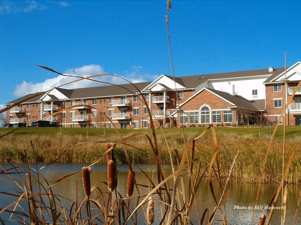 A large brick senior living apartment building with multiple balconies, set behind a pond with tall reeds and cattails in the foreground under a clear blue sky.