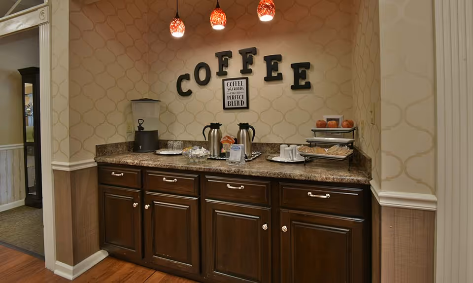 Small coffee station with dark wood cabinets, coffee urns, pastries and 'COFFEE' letters on the wall.