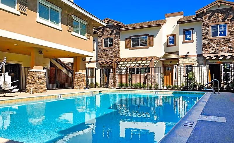 Outdoor swimming pool with clear blue water surrounded by a fence, adjacent to a multi-story residential building with stone and stucco exterior under a bright blue sky.