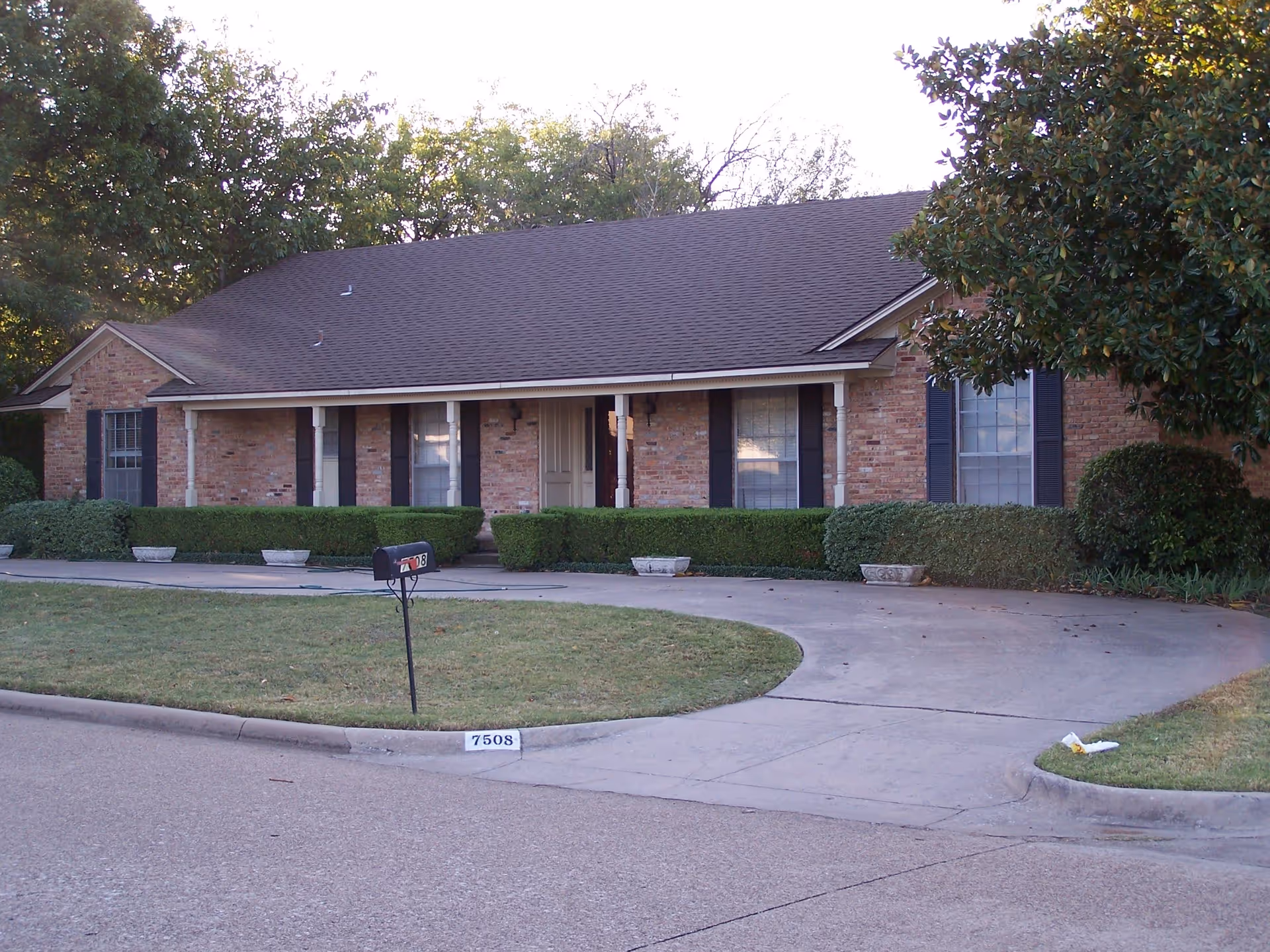 Single-story brick house with a dark shingled roof, white columns on the front porch, black shutters on the windows, neatly trimmed bushes along the front, a curved concrete driveway, a black mailbox on a post, and a house number 7508 painted on the curb.