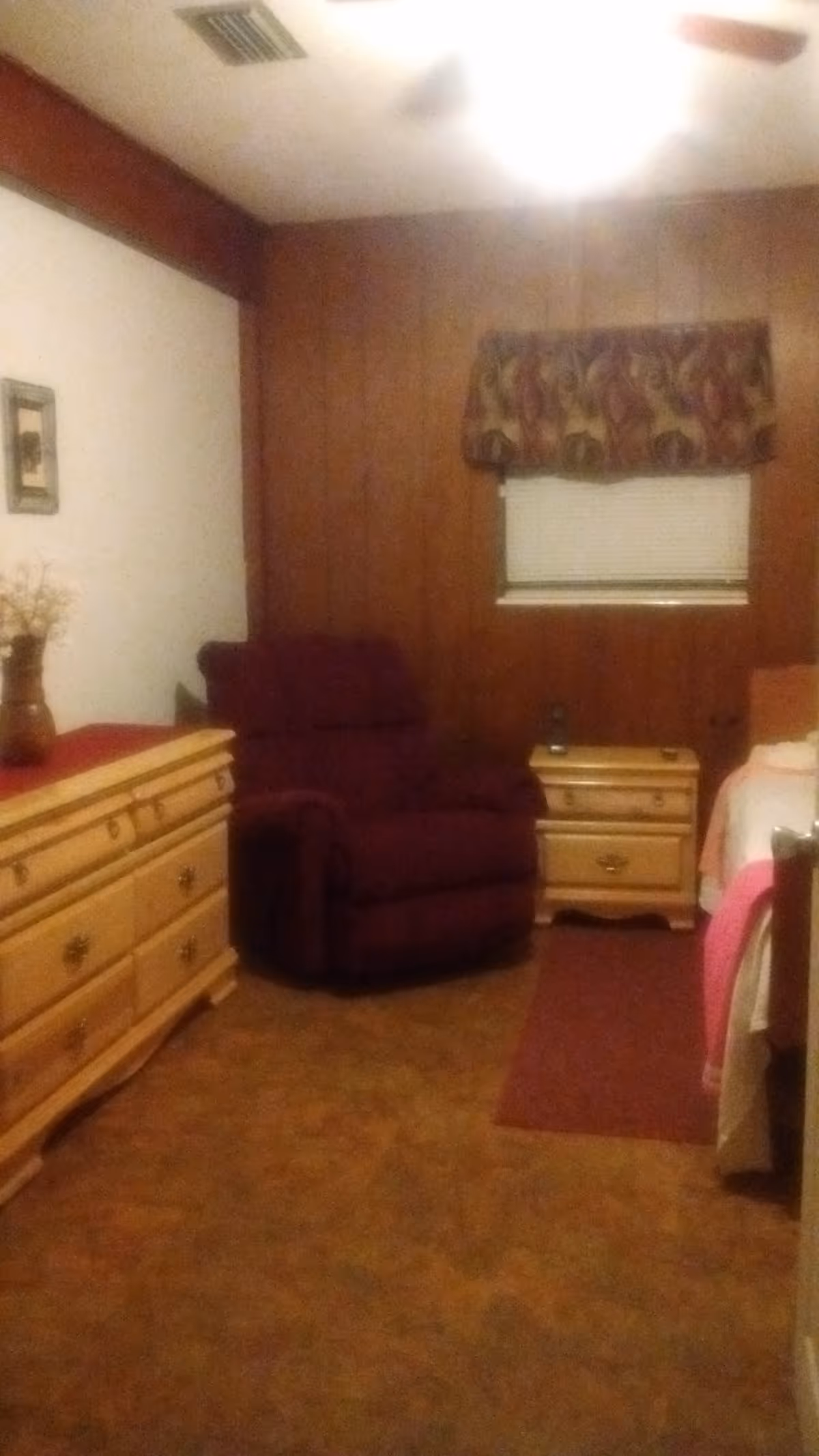 Small bedroom with wood-paneled walls, a burgundy recliner, light wood dresser and nightstand, and a bed partially visible on the right.